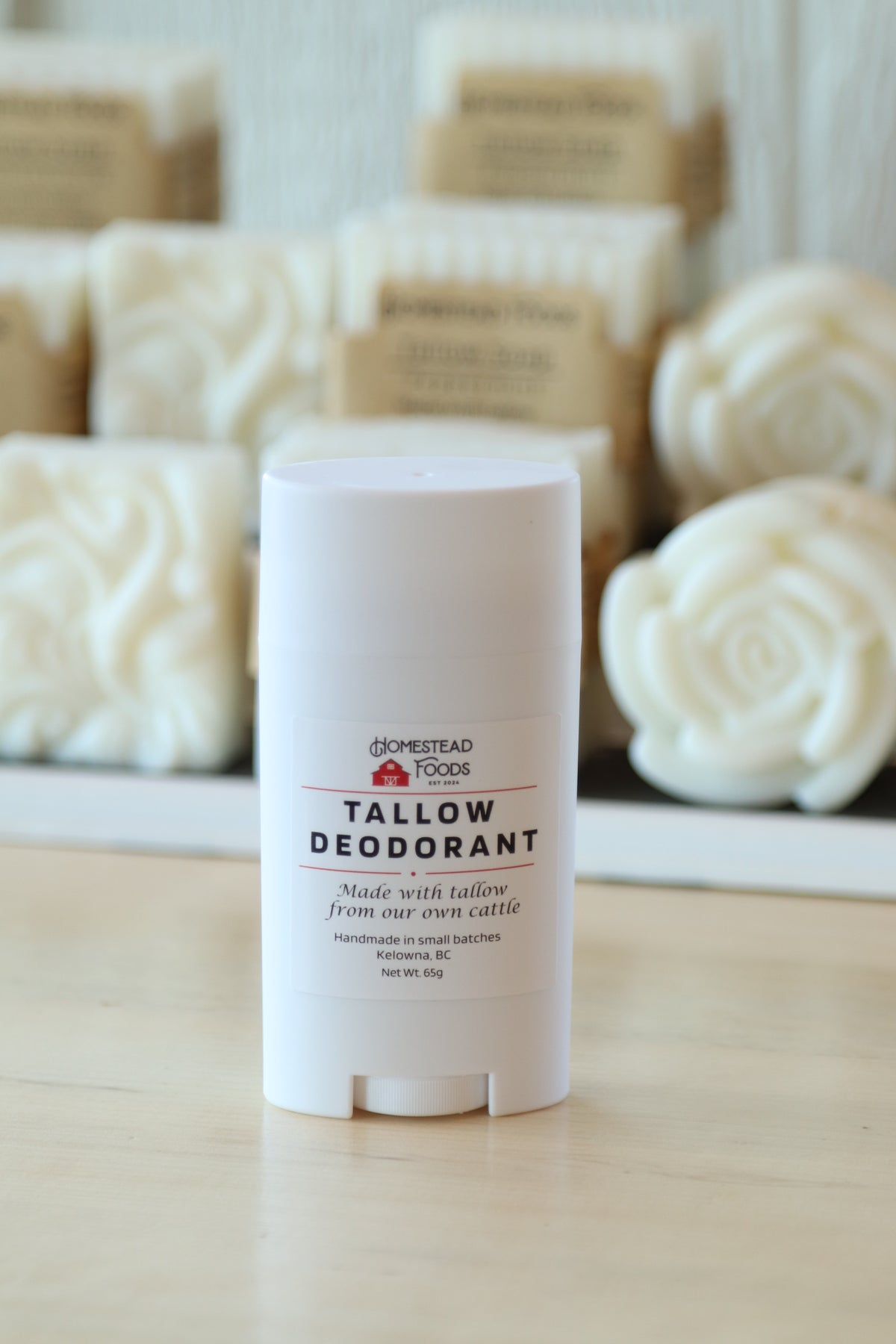 A white plastic stick container of Homestead Tallow Deodorant standing on a light wood surface, with blurred bars of soap in the background.
