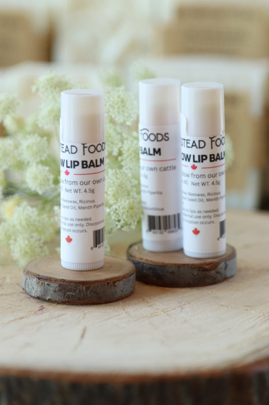 Three white tubes of Homestead Tallow lip balm arranged on small round wooden slices with blurred pale flowers in the background.