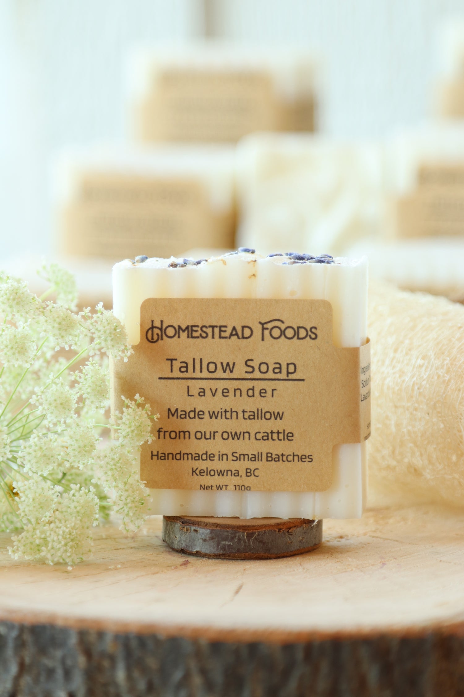 A close-up of a bar of Homestead TTallow Soap labeled Lavender, wrapped with a tan paper label, resting on a wooden surface with blurred soap bars in the background.